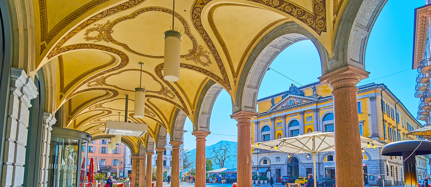 LUGANO, SWITZERLAND - MARCH 25, 2022: The scenic arcade of historic building on Piazza della Riforma square opens the view on the square, outdoor cafes and Palazzo Civico (Town Hall), on March 25 in Lugano